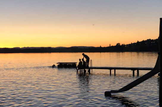 Menschen Baden im See und sitzen auf einem Steg bei Sonnenuntergang