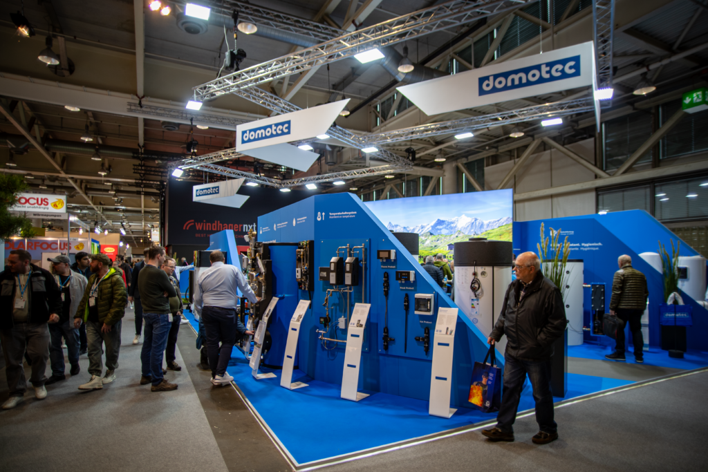 Auf einem Messestand von Domotec sind verschiedene Heizungs- und Sanitärgeräte auf blauen Ständern ausgestellt. Mehrere Personen beobachten die Exponate, und der Bereich ist mit Oberlichtern und Bannern gut beleuchtet.