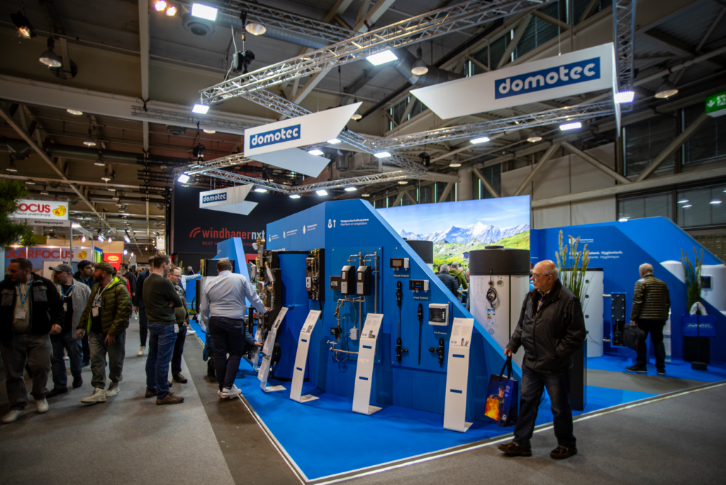 Un groupe de personnes visite le stand bleu et blanc de Domotec lors d'un salon professionnel, observant divers appareils et équipements techniques exposés sous une signalétique et un éclairage lumineux suspendus.